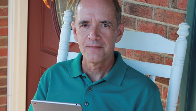 Photo of an adult man sitting on a rocking chair holding a tablet. 