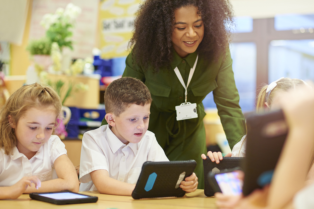 four primary aged schoolchildren sit at their desks and look engrossed in the work that they are doing on their digital tablets. Their schoolteacher is looking over their shoulder and joining in with pupils. They are all wearing white school shirts