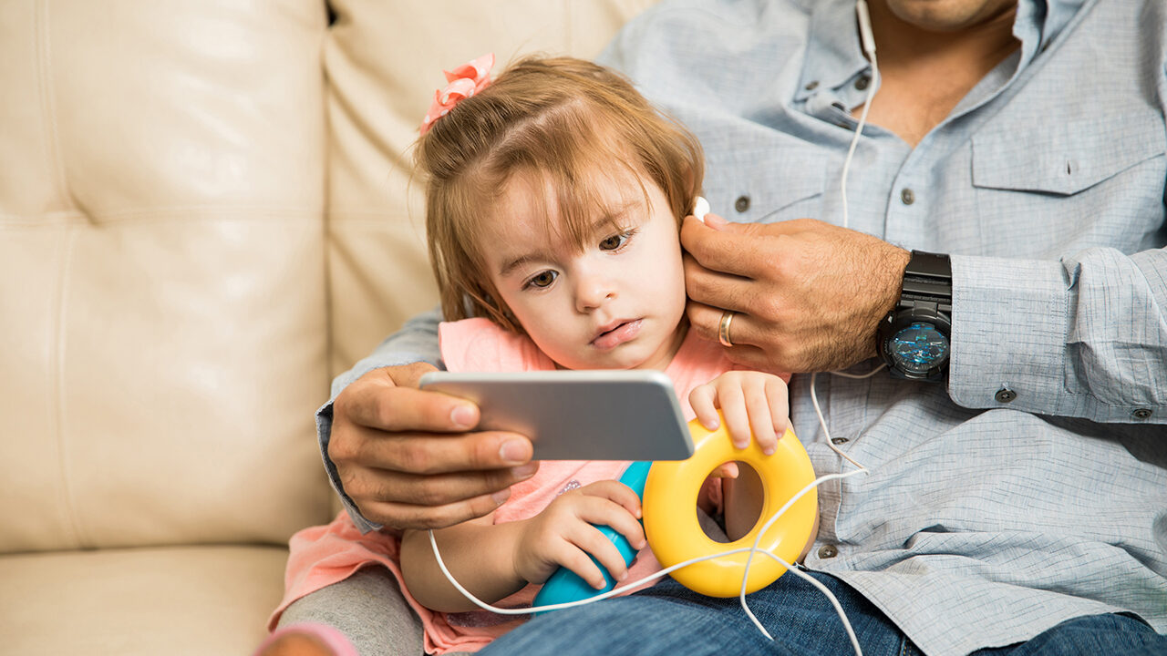 Toddler sitting with an adult on the couch listening to a story from a mobile device